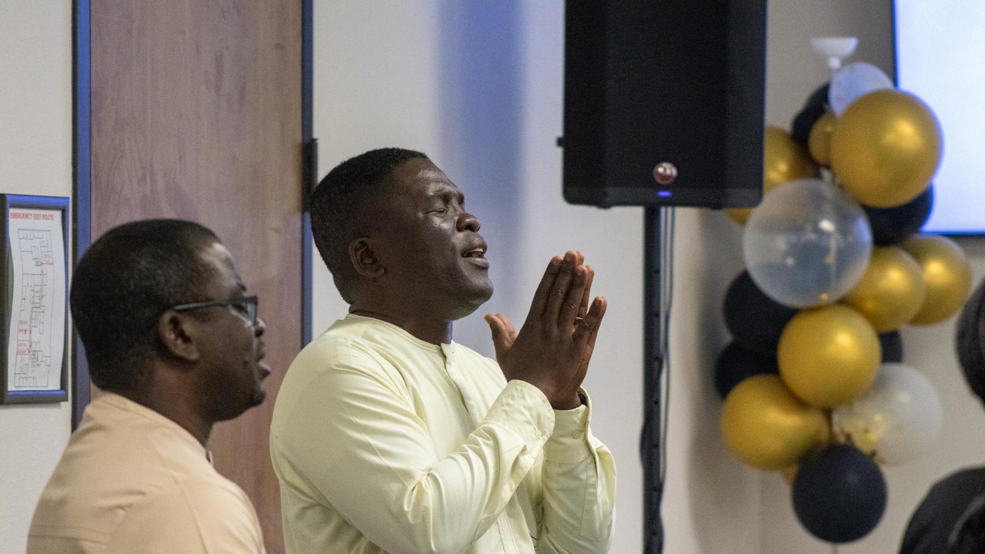 Two men standing side by side with hands raised in prayer.